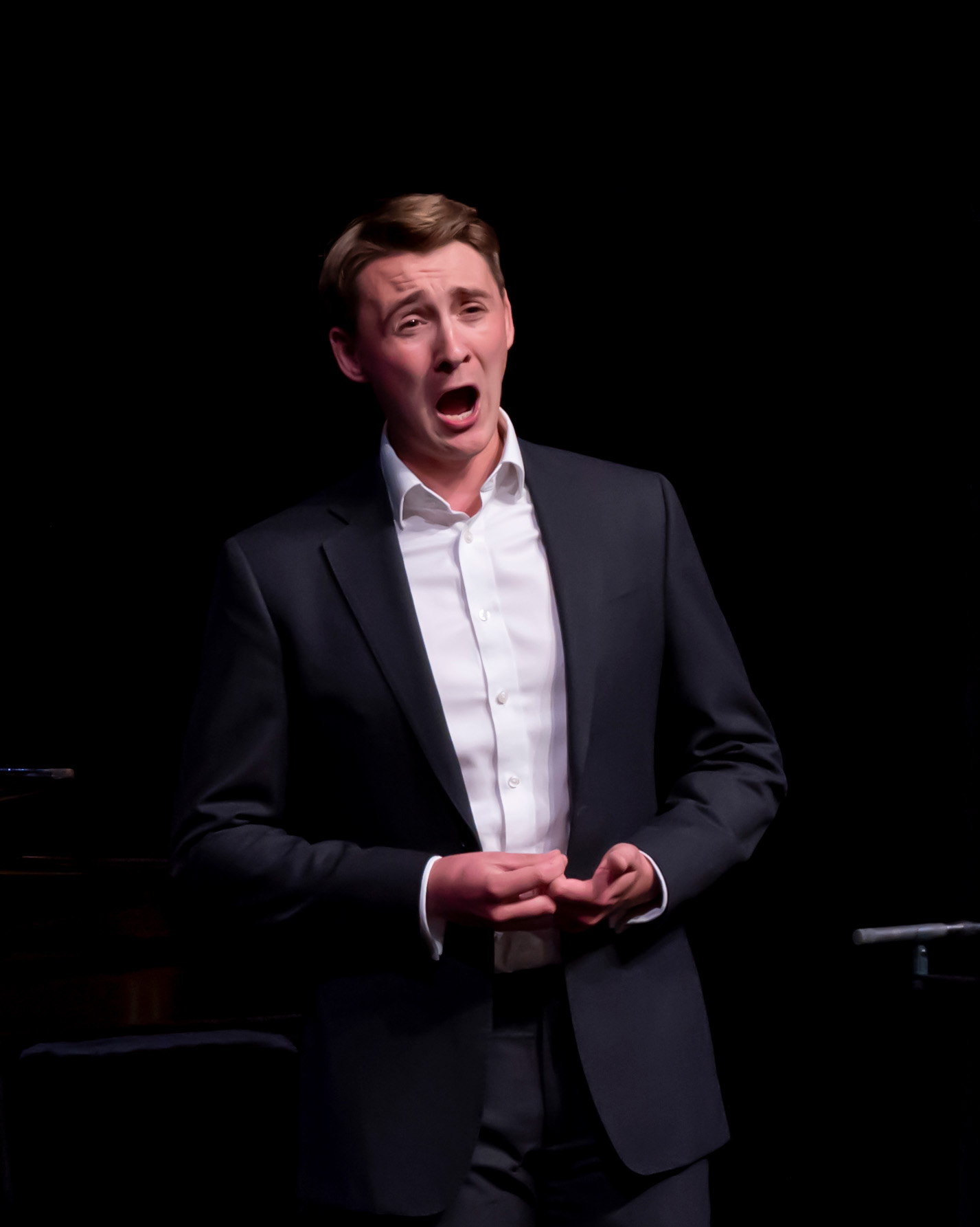 A man wearing a suit, singing on stage, lit by a spotlight.